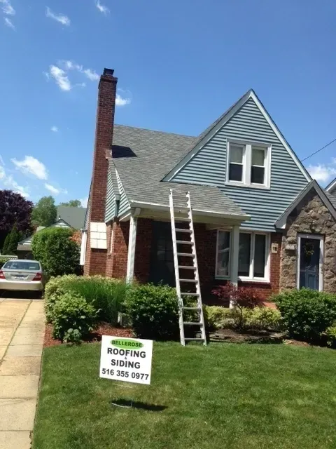 House with roofers’ ladder and sign in front lawn, brick chimney, gray siding, sunny day