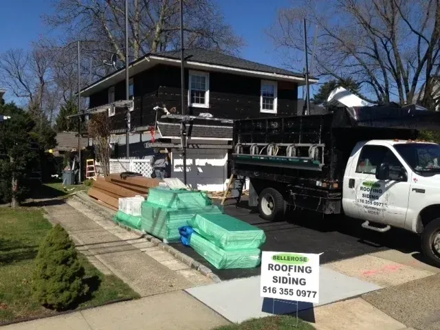 Roofing materials and trucks outside a house under repair, with a sign advertising roofing and siding.