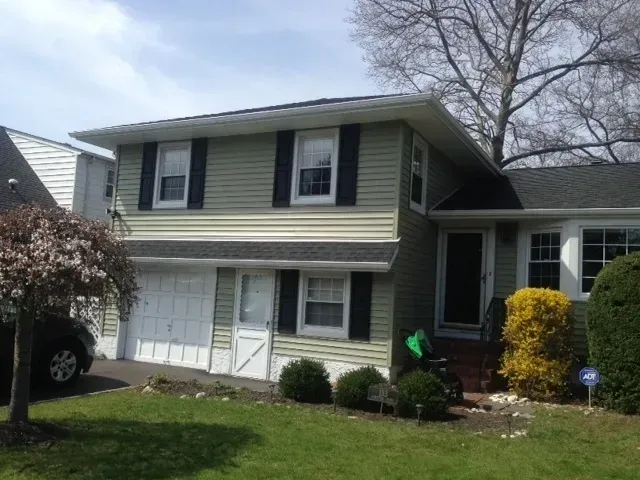 Two-story suburban house with beige siding, white garage doors, and a front yard with shrubs and a tree.