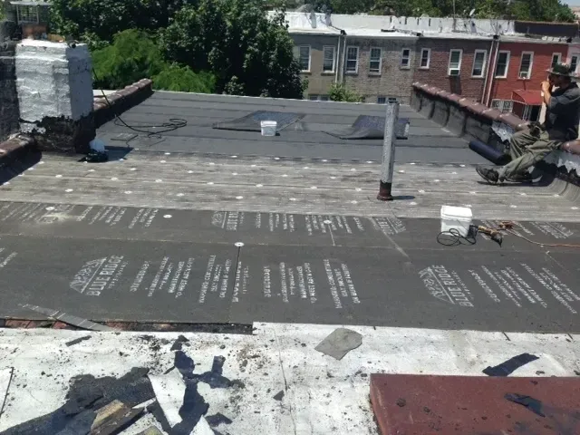 Rooftop with black tar surface, white vent pipes, and neighboring buildings in the background