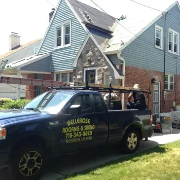 Roofing company truck parked outside a house under repair, with ladders and workers on the roof.