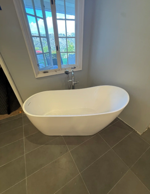 Modern bathroom with a white freestanding bathtub under a window on gray tile floor.