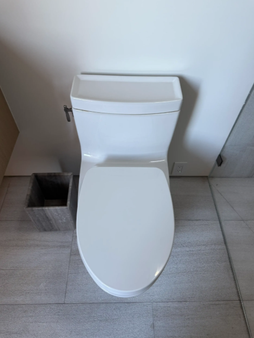 White toilet in a tiled bathroom with a small trash bin beside it.