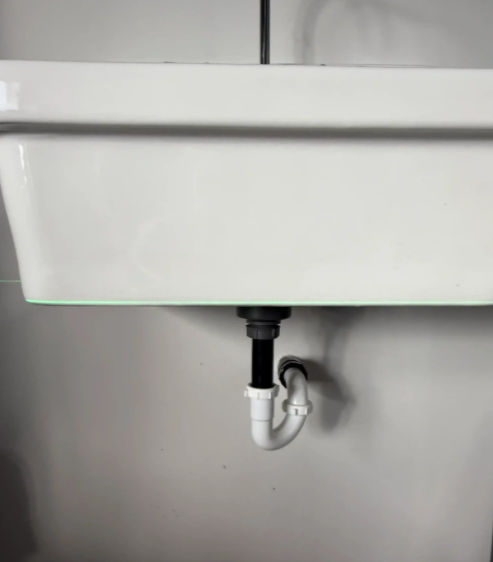White wall-mounted sink with exposed drain pipe and trap against a gray wall.