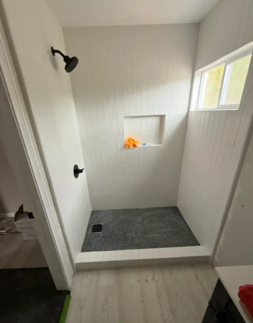 Small white tiled shower stall with black showerhead, gray floor mat, and window opening with orange item inside