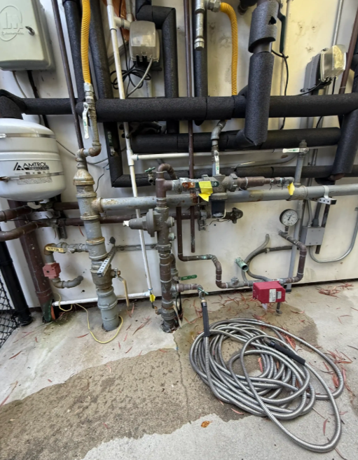 Exposed plumbing and valves in a utility room with coiled hose on the floor.