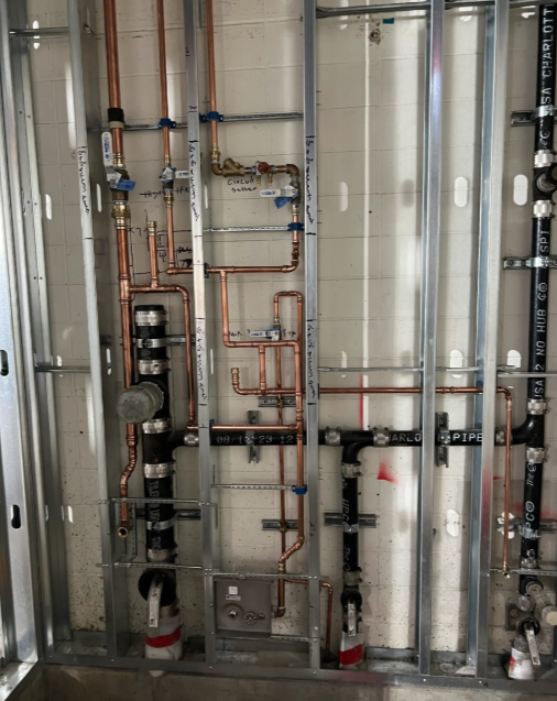 Mechanical room with exposed copper pipes, valves, and insulated tanks behind a wall frame.