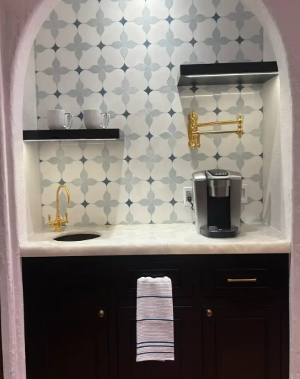 Small kitchen nook with patterned tile backsplash, dark cabinets, sink, shelves, and a coffee maker.