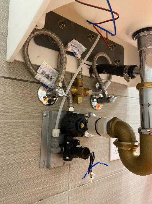 Under-sink plumbing with pipes, valves, and flexible hoses mounted against tiled wall.