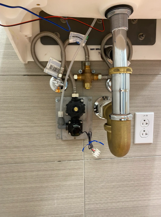 Under-sink plumbing and valves with exposed pipes, wiring, and a wall outlet in a tiled utility area
