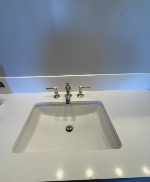 White bathroom sink with chrome faucet and handles on a clean countertop against a light wall.
