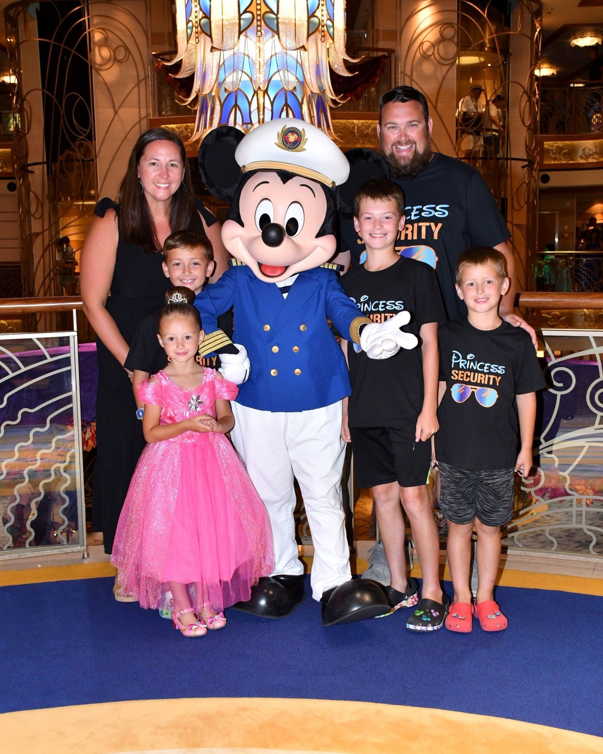 Family posing with Mickey Mouse on a cruise ship. Adults, children, and Mickey are smiling, posing for a photo.