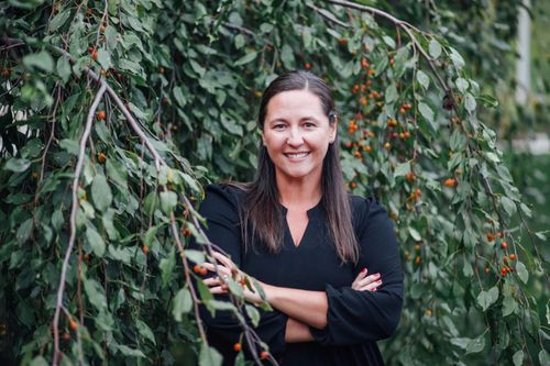 Woman on beach with arms outstretched, wearing white top, blue patterned skirt, sunset background.