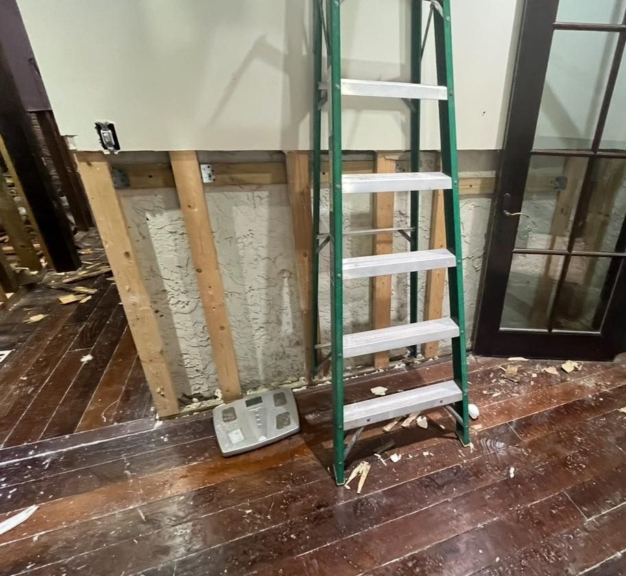 Green ladder leans against a partially demolished wall in a room with hardwood floors. A scale sits on the floor.