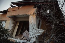 Damaged building facade, roof collapsed, curtains torn, exterior view, bare tree branches.