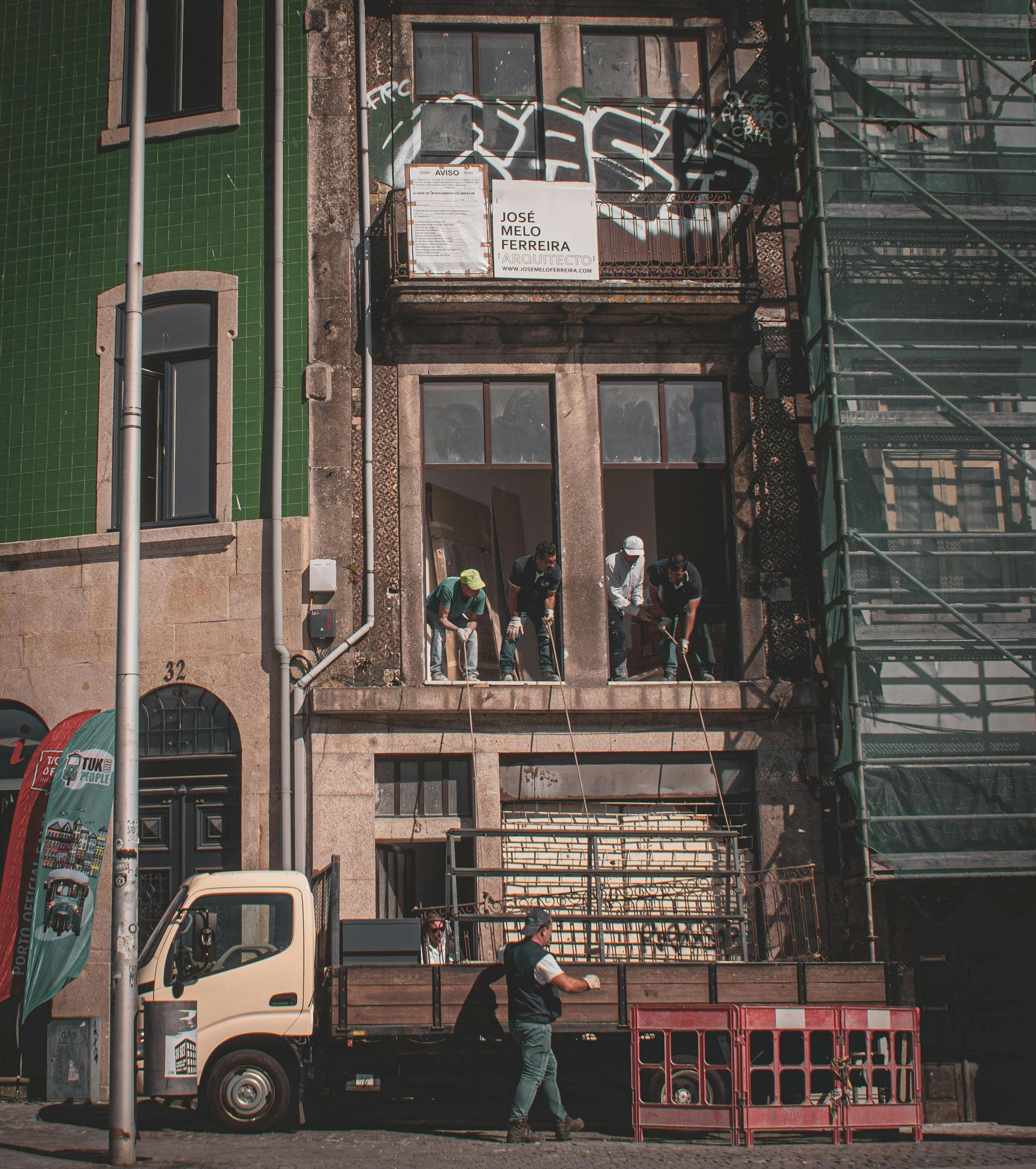 Workers on a truck load goods beneath a building with graffiti, partially covered in scaffolding.