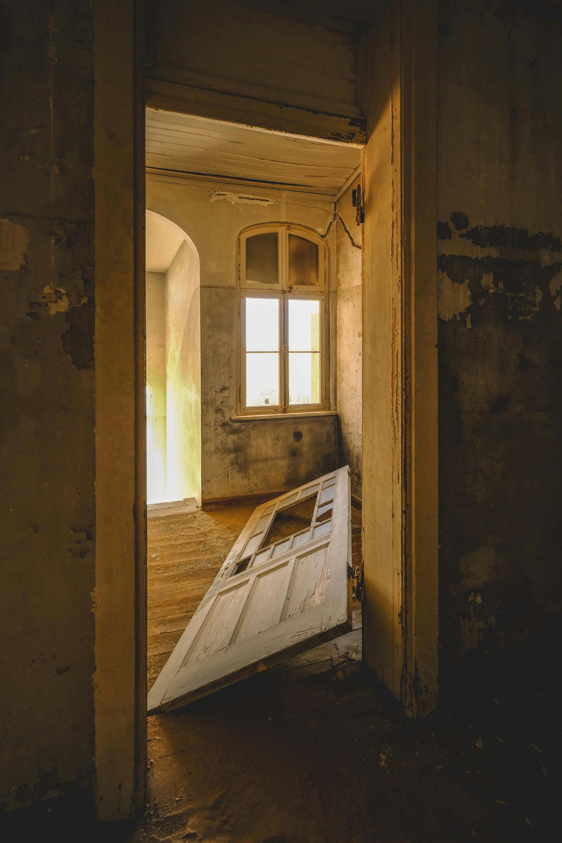 Interior shot of abandoned room with broken door and window. Dilapidated walls, light streams through the window.