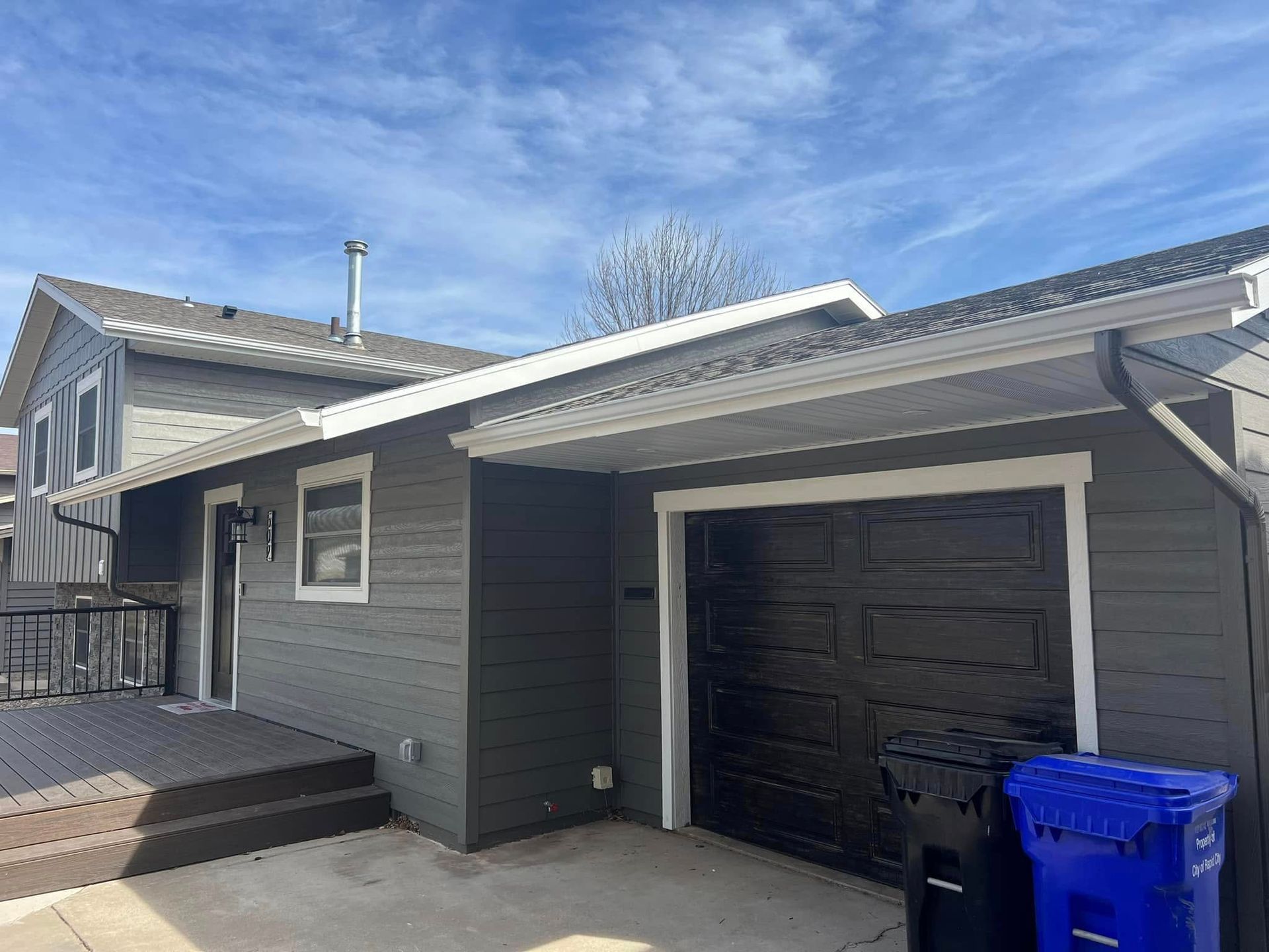 Grey house with black garage door, wooden deck, and blue trash bin. Sunny day with blue sky.