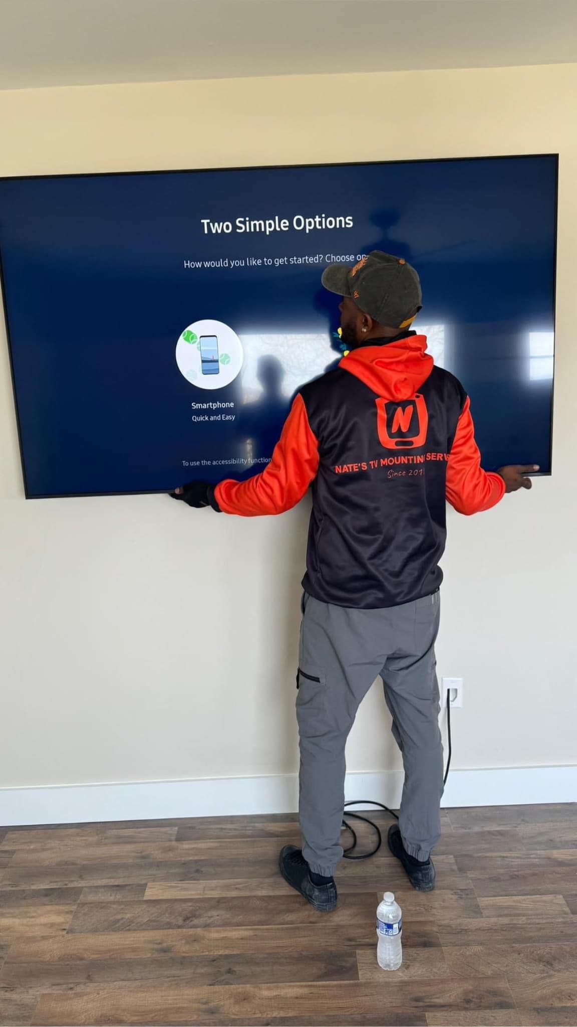 Person holding a large TV against a wall, about to install it. They wear a red and black jacket and gray pants.