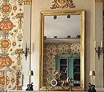 Large gold-framed mirror reflects ornate wallpaper and turquoise cabinet; flanked by sconces and lamps.