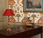 Red lamp on a wooden desk; floral wallpaper and framed art.