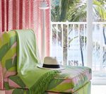 Chaise lounge with green and pink pattern, hat, blanket, palm trees visible through window.