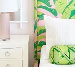 White nightstand with pink lamp next to a bed with a floral patterned headboard and a decorative green pillow.