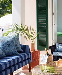Blue striped couch, green shutter, palm frond, orange stool on a white veranda.