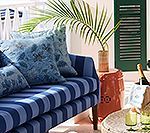 Blue striped sofa with patterned pillows, palm frond, orange table, green shutters.