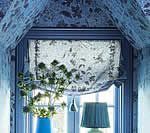 Window with floral roman shade, blue decor, vase with greenery, and lamp.
