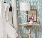White lamp and flowers on a small table next to a bed with white bedding, teal patterned wallpaper.