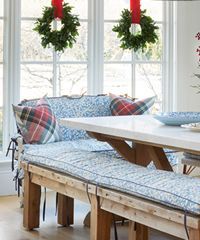 Wooden bench with blue patterned cushions, plaid pillows, set near a window with Christmas wreaths.