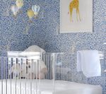 A nursery with a clear crib, blue patterned wallpaper, and giraffe artwork. Decorative balloons hang above the crib.