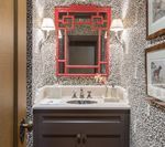 Red mirror hangs above vanity in a bathroom with a patterned wall and sconces.