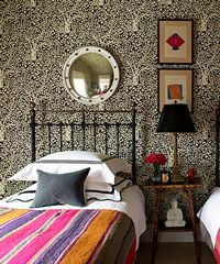 Bedroom with patterned wallpaper, metal bed, round mirror, side table with lamp, and two framed artworks.