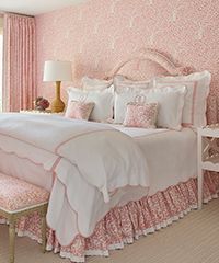 Pink bedroom with floral wallpaper, bed with ruffled bedding, and a side table.