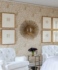 White tufted chairs in a bedroom with floral wallpaper, gold framed artwork, and a sunburst mirror.