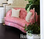 Pink patterned settee with two cream pillows, beside a potted plant, indoors.