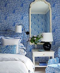 Bedroom with blue patterned wallpaper, white bedding, ornate mirror, side table, lamp.