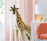 Giraffe standing near a window with pink patterned curtains, a lamp, and an orange chair.