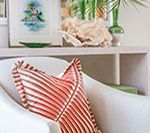 White armchair with orange-striped pillow; shelf with coral, green vase, and artwork.