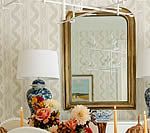 Gold-framed mirror reflects a floral centerpiece and lamps on a table in a room with patterned wallpaper.
