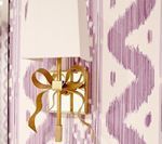 Gold bow-shaped wall sconce with white lampshade against a patterned purple and white wall.