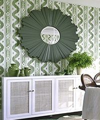 Green sunburst mirror above a white cabinet with woven doors, against green patterned wallpaper.
