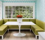 Green booth seating around a white table in a light-filled room with large window and patterned blue wallpaper.