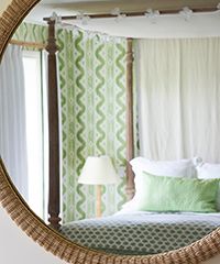 Round mirror reflecting a bedroom with a bed, green accents, and patterned curtains.