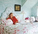 Woman in red top and white pants relaxes on floral bed in a blue-walled bedroom.