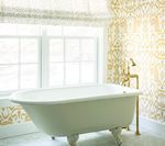 White clawfoot bathtub in a light-filled bathroom with gold patterned wallpaper and fixtures.