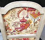Chair back with Asian-themed fabric. Cream-colored frame. Fabric features a pagoda, trees, and other stylized elements in red, gold, and green.