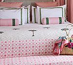 Bed with pink and white bedding, decorative pillows, and a straw bag.